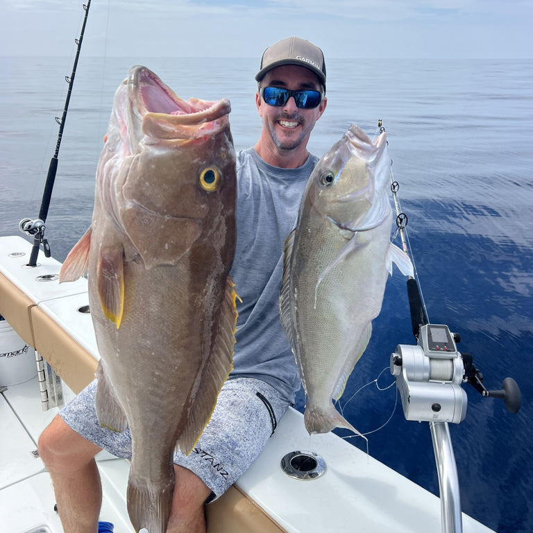 Man on a boat holding two large fish with a fishing rod and reel in the background.