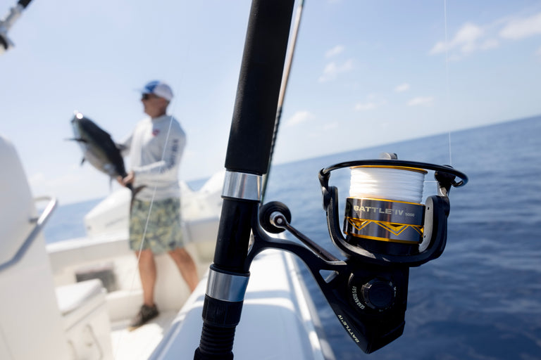 Penn Battle IV Spinning Combo in a boat holder over the ocean with a man in the background holding his prized catch of the day.