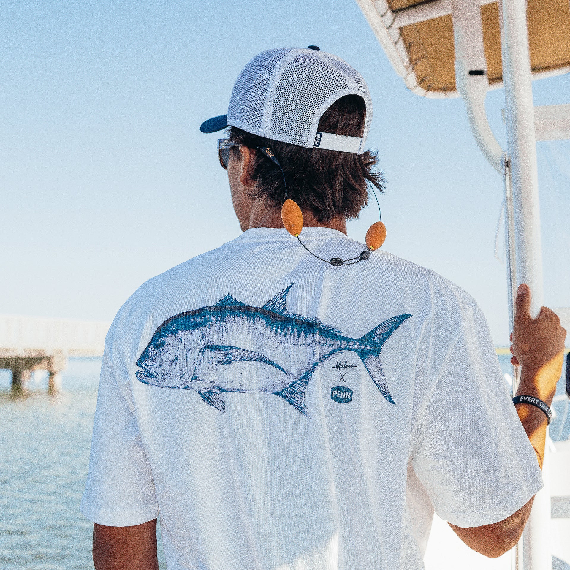 Person on a boat wearing a white shirt with a fish design and a cap, looking out over water.