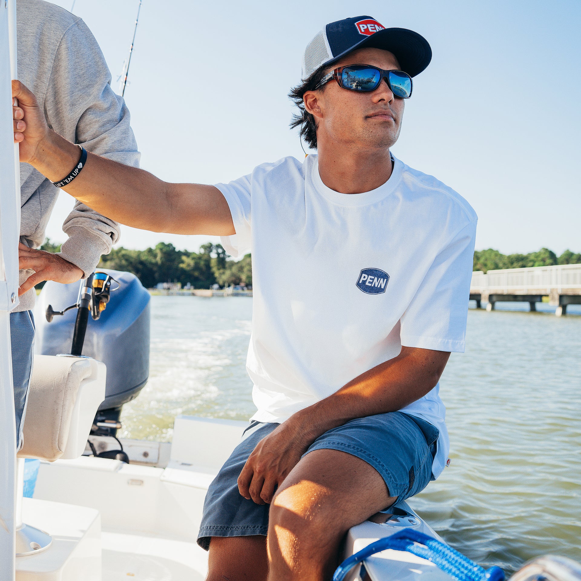 Man on a boat wearing a white t-shirt with a brand logo, sunglasses, and a cap.