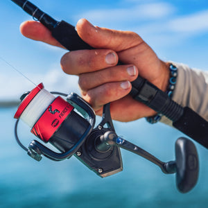 A close-up lifestyle shot of a person's hand holding a spinning fishing rod with a red and black Reelfish brand spool, set against a blurred background of blue sky and water.