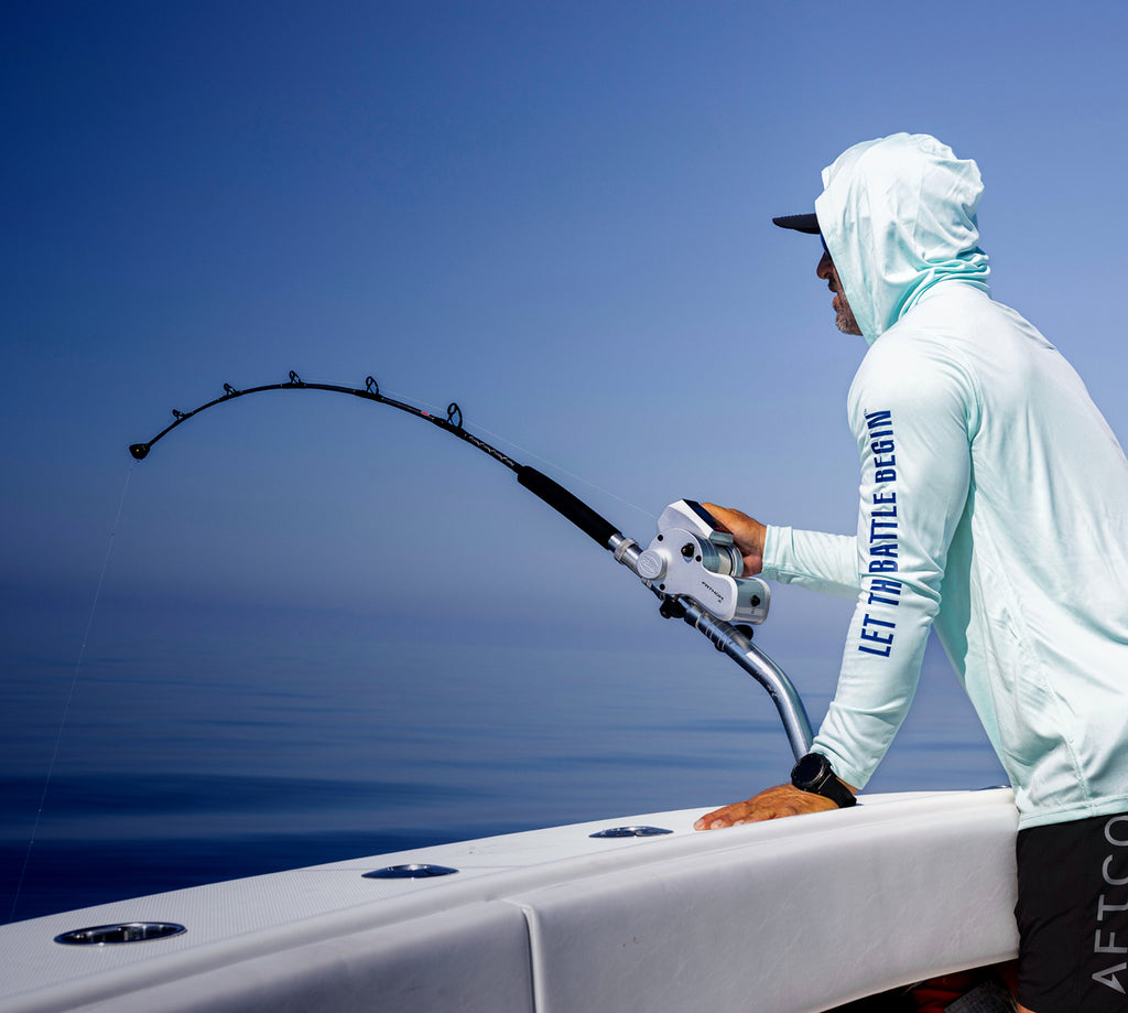 Person on a boat with a large fishing rod, wearing a light-colored hoodie and cap, against a clear blue sky.